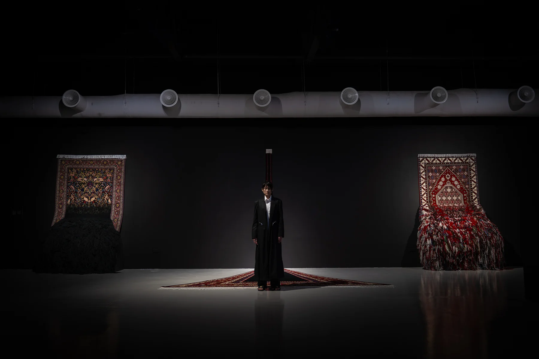 Figure standing between two hanging carpets in a dark gallery