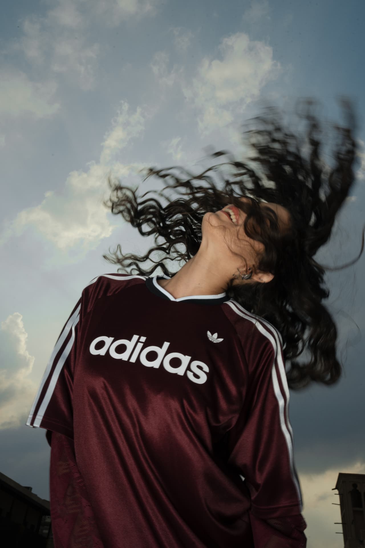 Woman tossing her hair in a maroon Adidas jersey against the sky