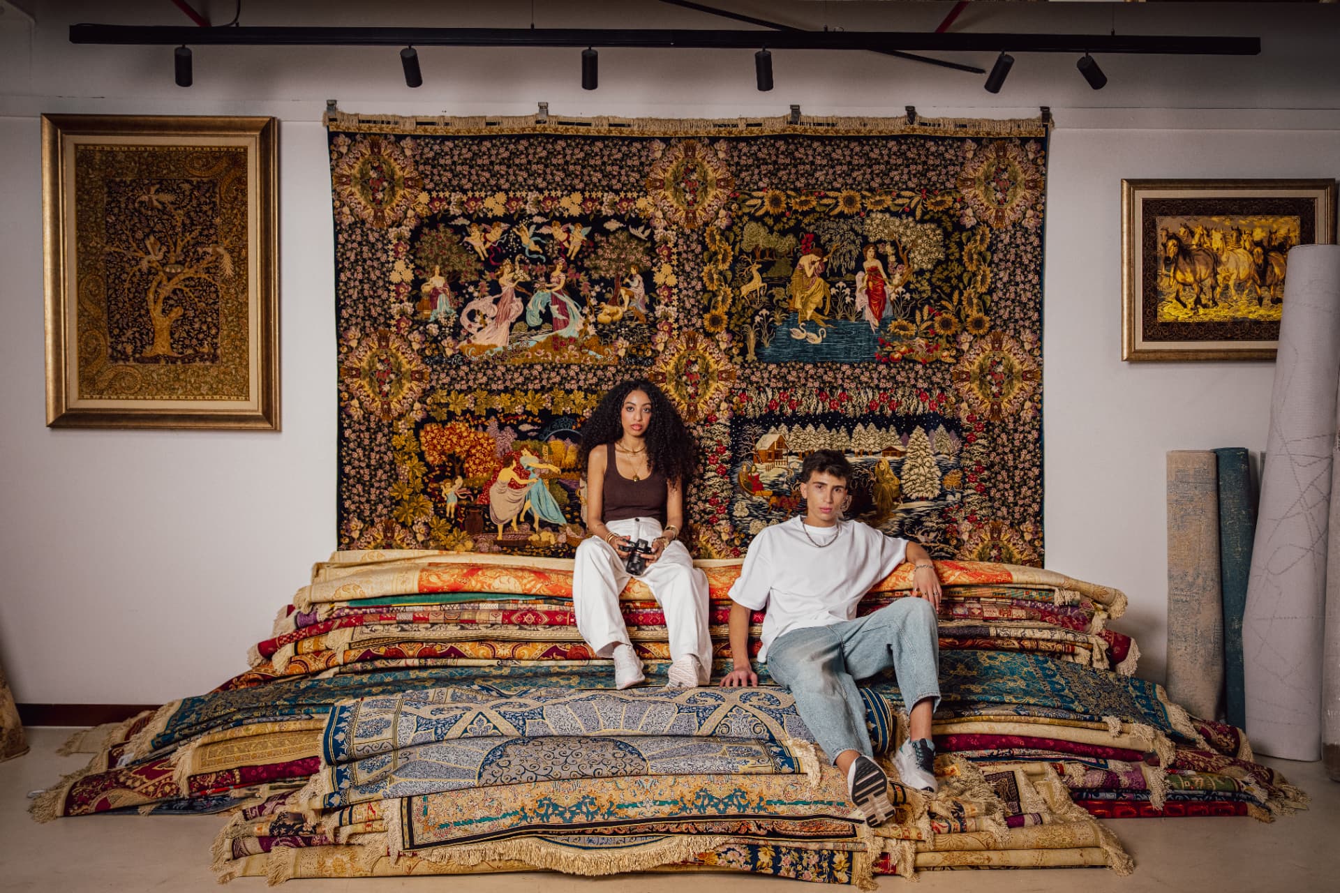 Two models seated on stacked carpets under a tapestry