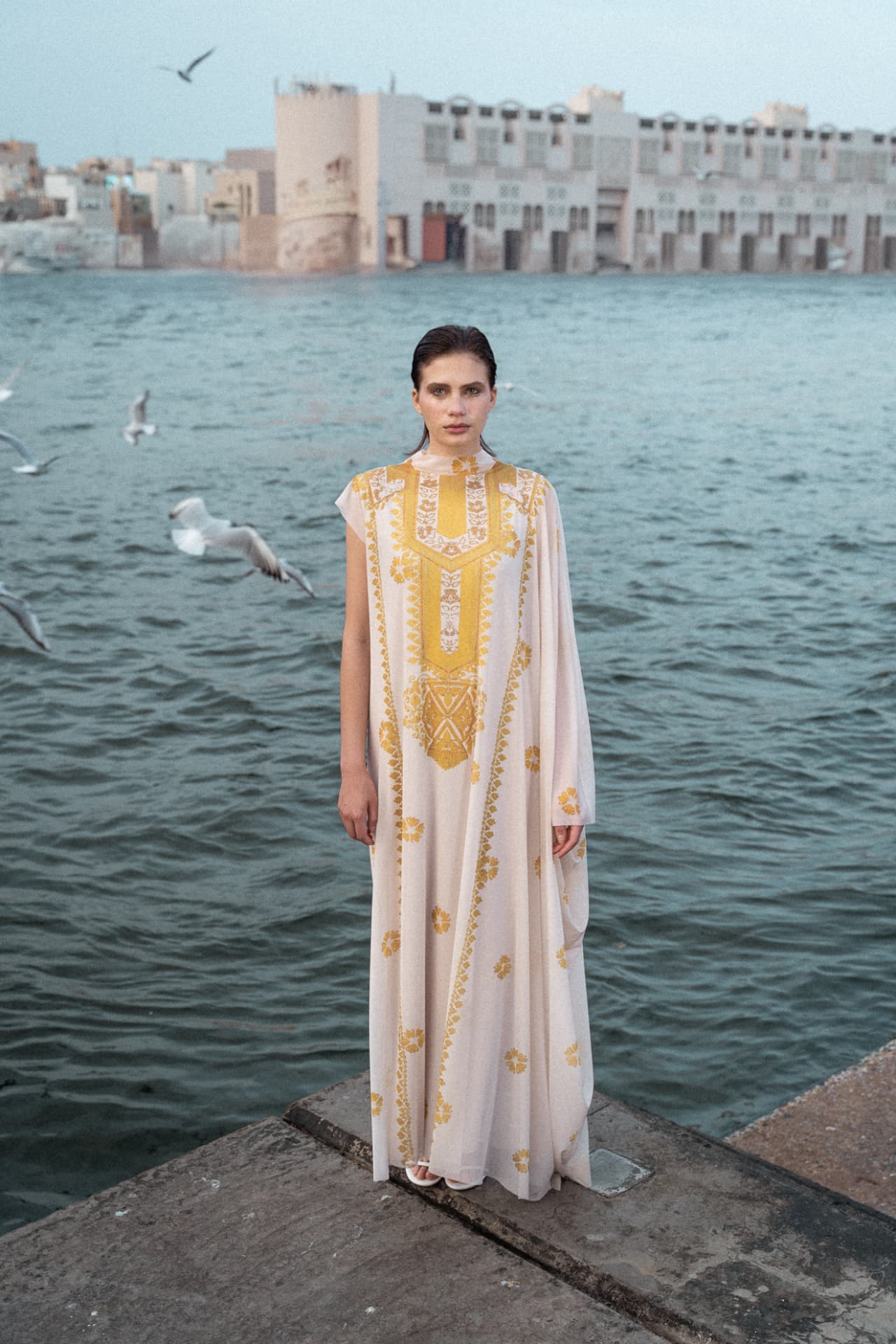 Model in white kaftan on the creek edge with gulls overhead