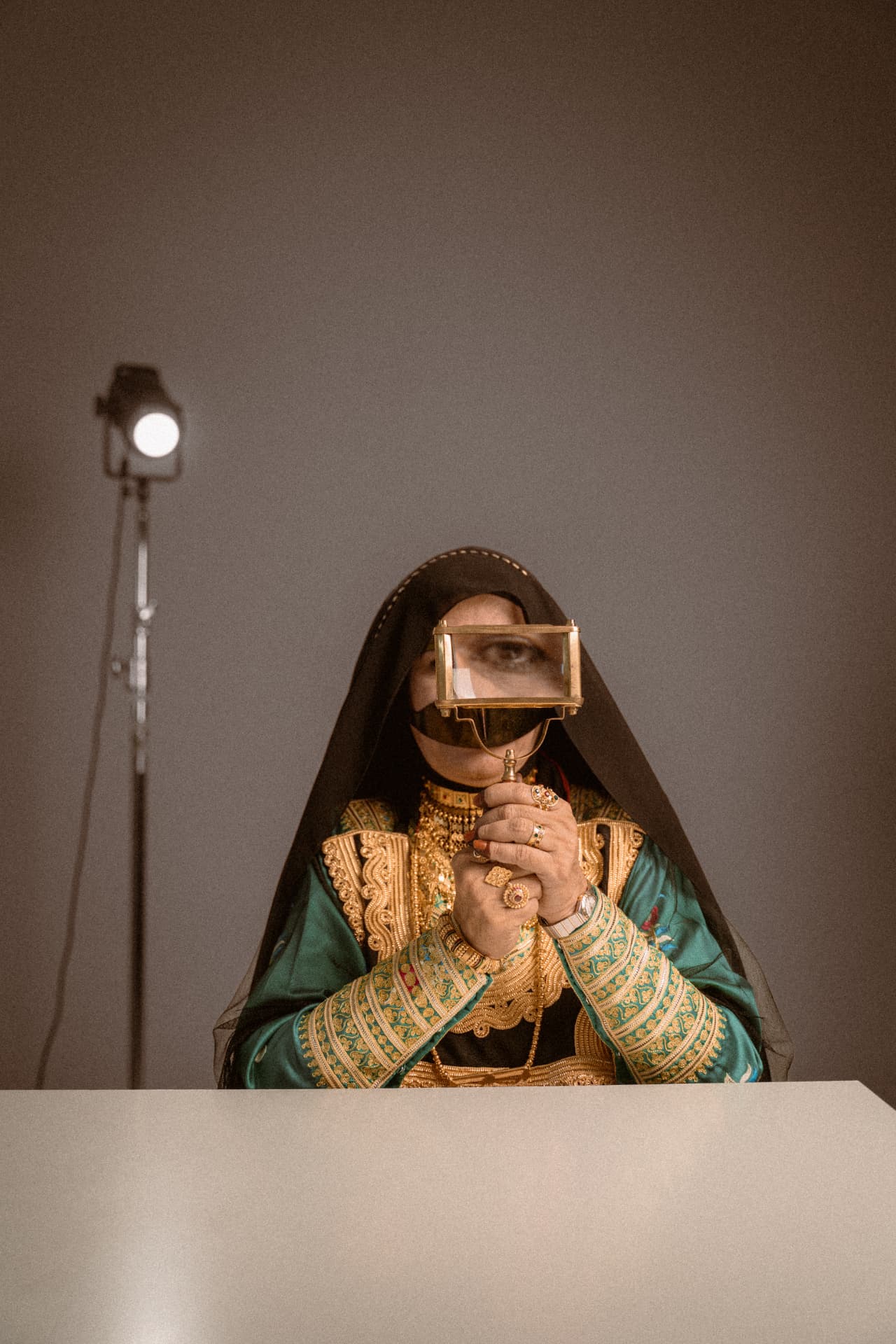 Woman in burqa holding a gold frame over her eyes under a studio light