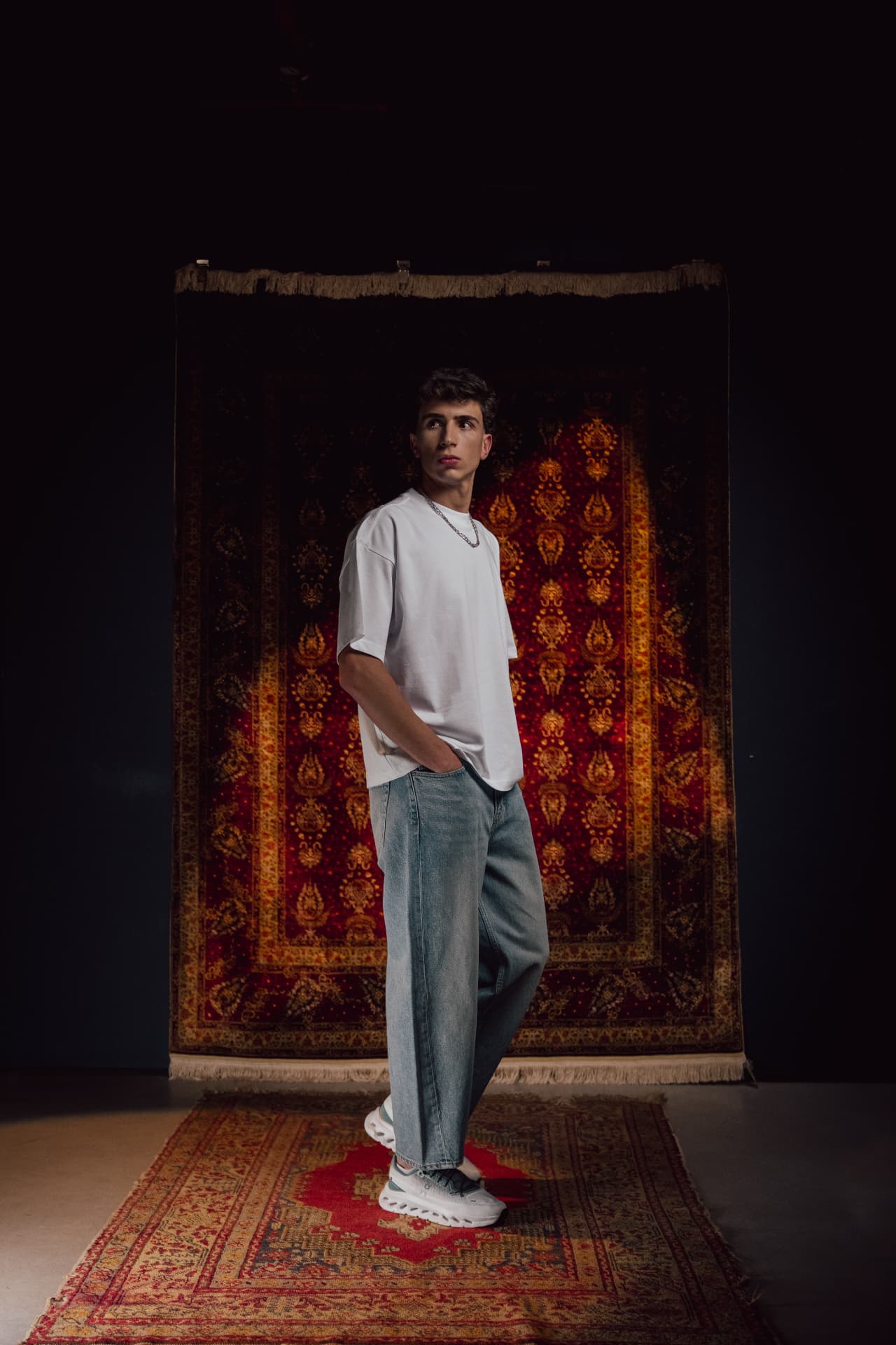 Model in white tee and jeans on a red rug before a hanging carpet