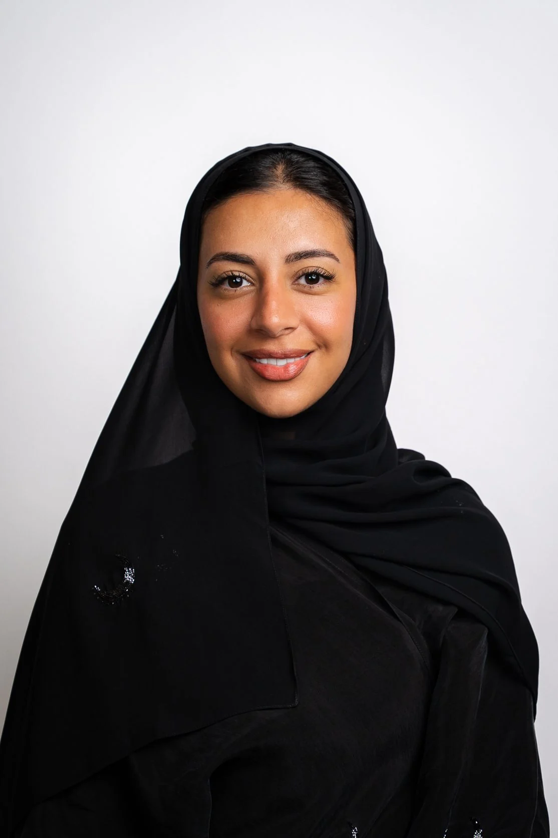 Smiling Emirati woman in a black abaya against a white backdrop