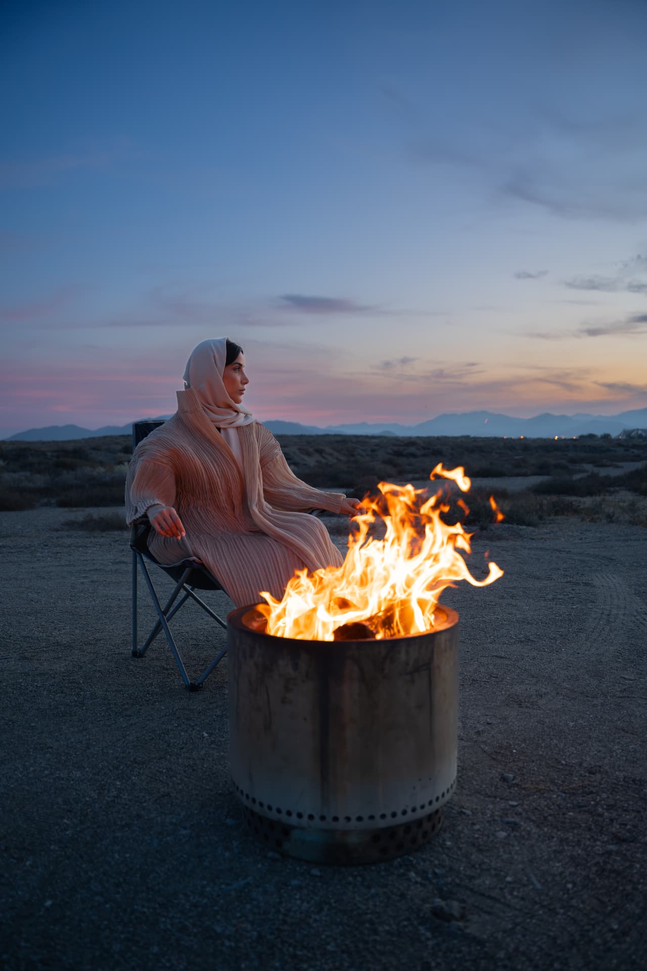 Woman in a headscarf beside a fire bowl at dusk in the mountains