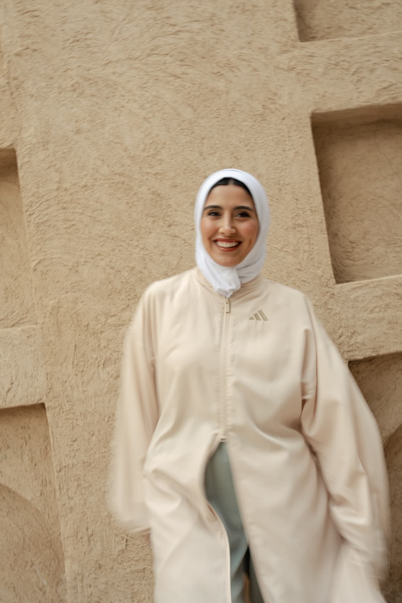 Smiling woman in a cream Adidas jacket against a sandstone wall