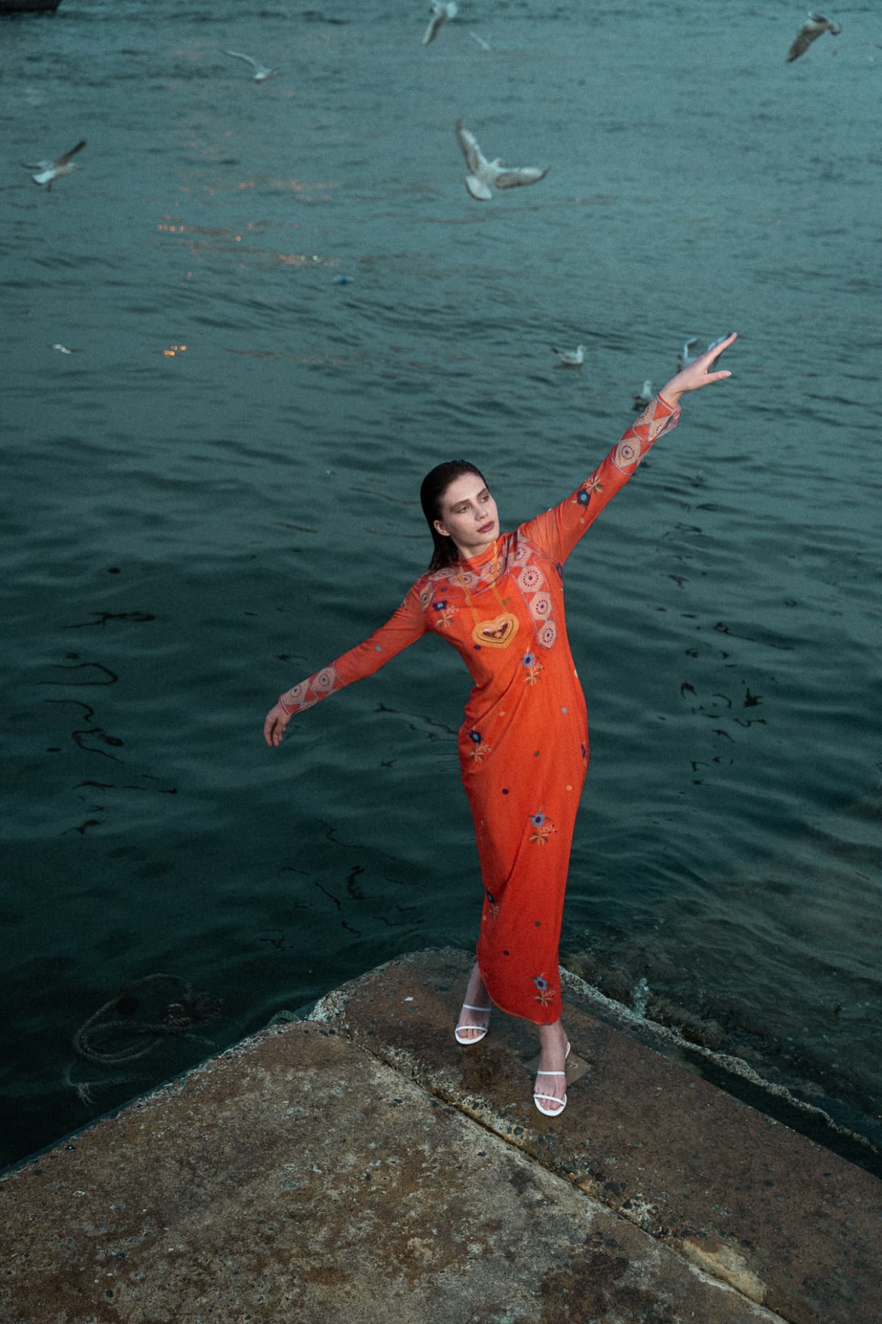 Model in orange dress reaching up on a creek-side jetty
