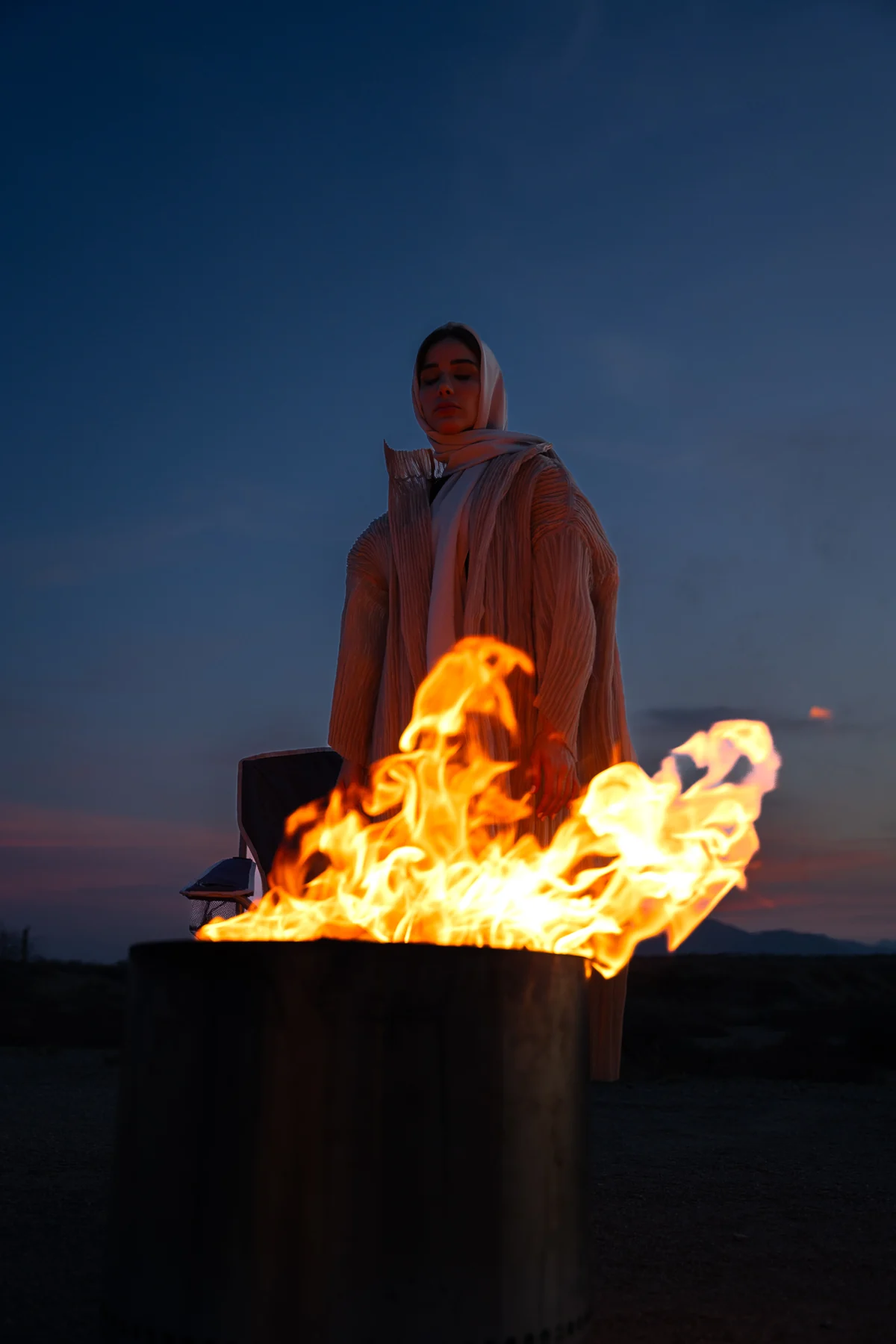 Woman in a pale headscarf beside a fire bowl at dusk